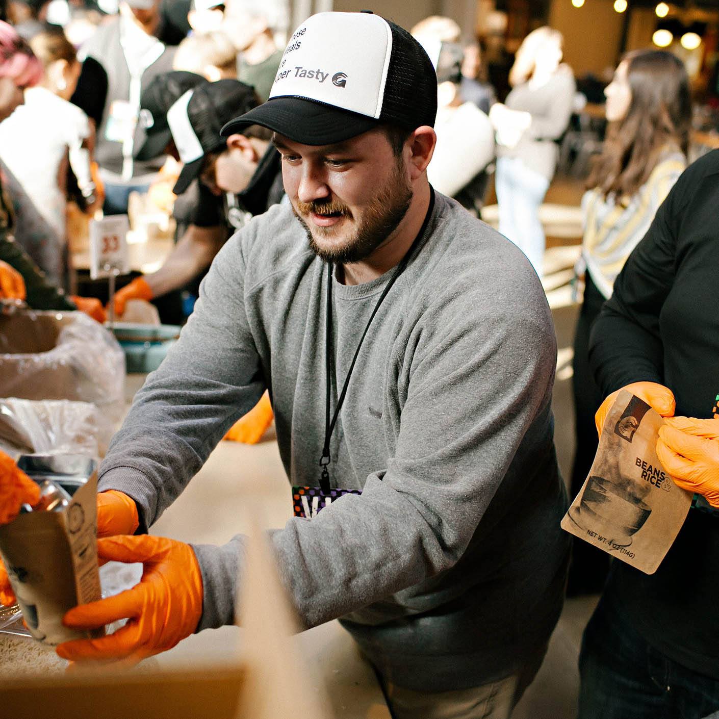 A man at a meal packing event hold a bag wearing gloves.